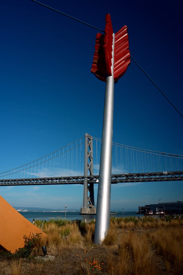 A large sculpture of an arrow is embedded in the ground near a bridge, with a clear blue sky in the background