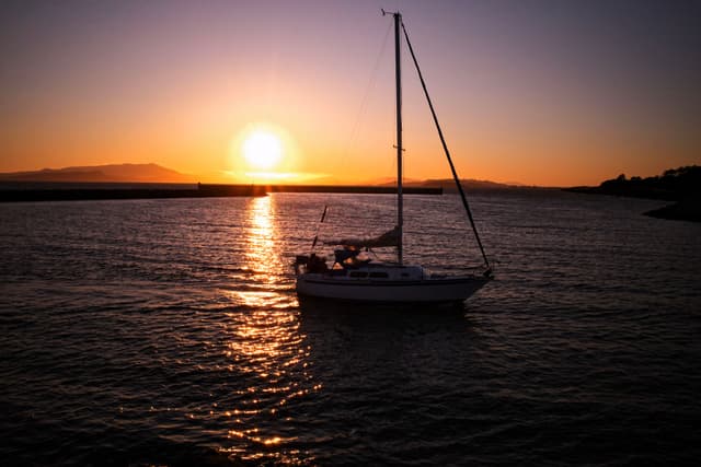 A sailboat on calm water during a vibrant sunset, with the sun low on the horizon and a silhouette of distant hills