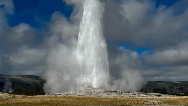 A powerful geyser erupts, sending a tall plume of water and steam into a cloudy sky over a rocky basin