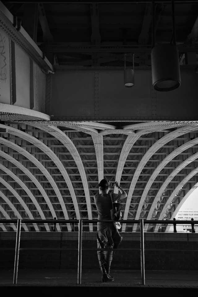A person standing on a platform, silhouetted against an architectural structure with curved beams, in black and white