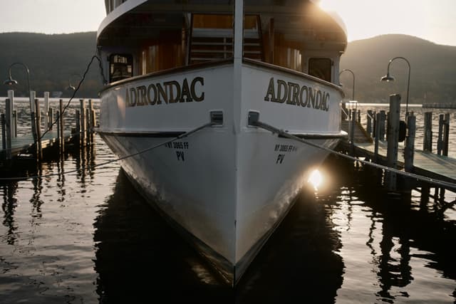 Bow-on view of a boat named “ADIRONDAC” docked between piers at sunset, reflections shimmering on calm water with hills in the background