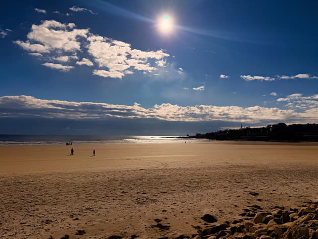 Wide sandy beach under bright sun with scattered clouds and a few distant walkers near the water