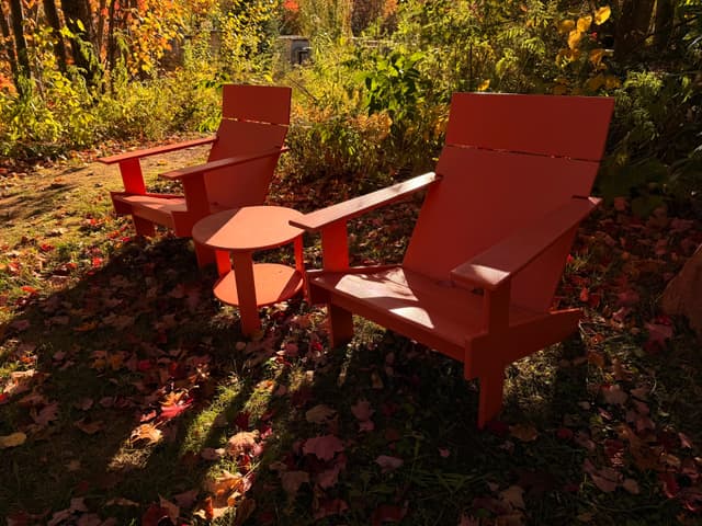Two red Adirondack chairs on a leafy lawn under autumn trees, dappled with sunlight