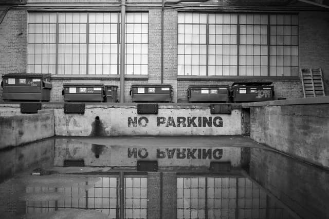 A row of train cars inside a building with large windows, reflected in a pool of water, and a NO PARKING sign on the wall
