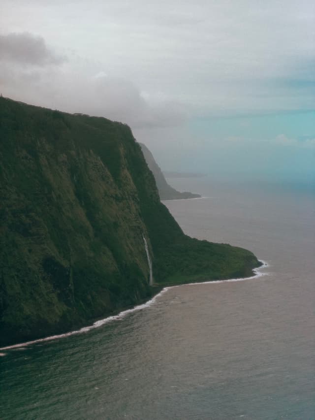Steep green cliffside along the coastline with a waterfall cascading into the sea and a distant horizon