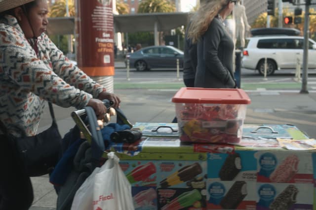 A street vendor pushes a cart covered with images of ice cream, carrying a red-lidded container and a bag, while a person walks by on the sidewalk