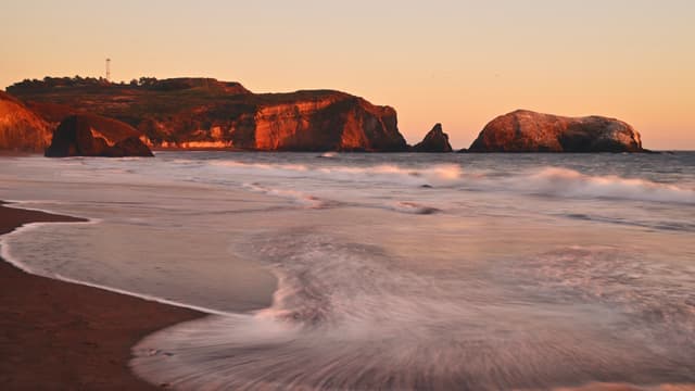 A serene coastal scene at sunset with gentle waves washing onto a sandy beach, rocky cliffs in the background, and a warm, golden glow illuminating the landscape