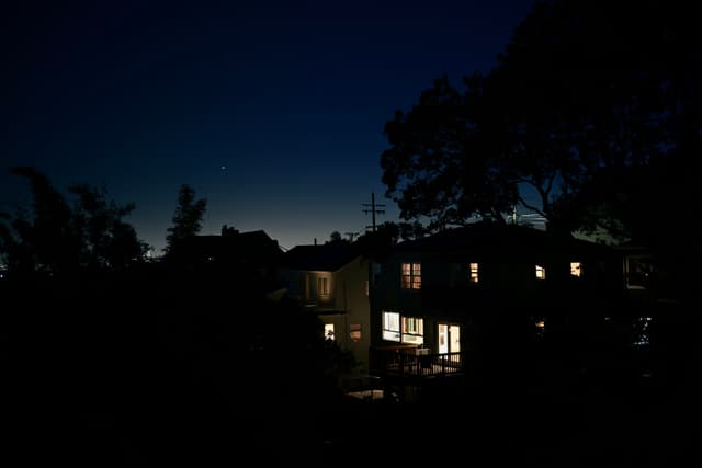 A nighttime scene with houses illuminated from within, surrounded by silhouetted trees against a dark blue sky