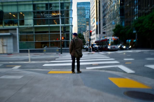 A person standing at a city intersection, surrounded by tall buildings and a bus approaching in the background