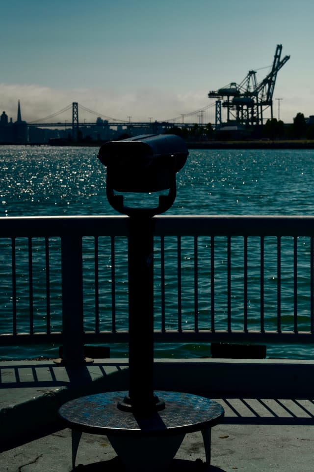 A waterfront scene with a coin-operated binocular viewer in the foreground, a body of water, a bridge, and cranes in the background