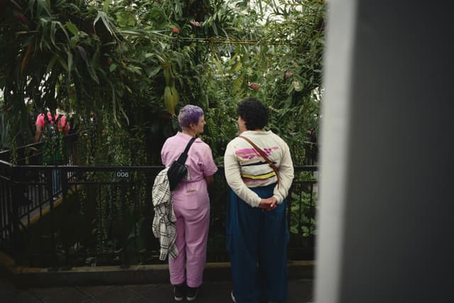 Two people standing on a balcony, surrounded by lush greenery, engaged in conversation