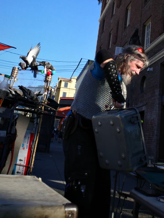 A person wearing a chainmail shirt and spiked arm guards is working with equipment on a street. A pigeon is flying nearby, and there are buildings and wires in the background