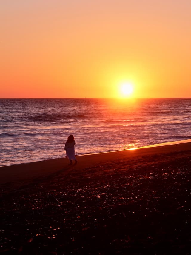 A person walking along a beach at sunset, with the sun low on the horizon and the sky filled with warm orange hues