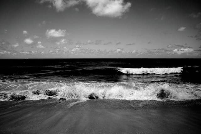 A black and white seascape with waves crashing onto the shore under a cloudy sky