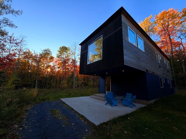 Modern dark-clad cabin with a cantilevered upper floor and large window, a small patio with blue Adirondack chairs, set in a wooded area with autumn foliage at sunset
