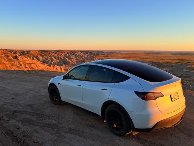 White Tesla parked on a dirt overlook at sunset, facing a vast rocky desert canyon
