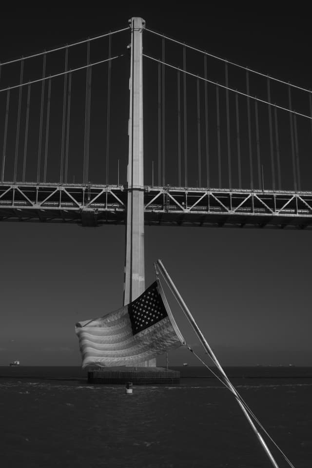 A black and white photograph of a suspension bridge with an American flag in the foreground