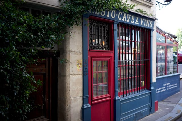 A quaint storefront with a red door and window frames, featuring the words BISTRO CAVE A VINS above. The building has a classic, rustic appearance with greenery on the side