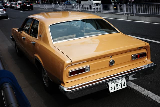 A yellow vintage car is parked on the side of a street, with a visible license plate and other vehicles in the background