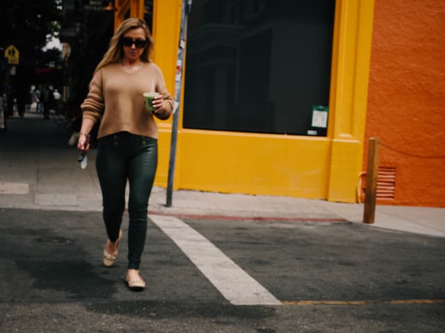 A woman walking on a street holding a drink, wearing sunglasses and casual clothing, with a bright orange building in the background