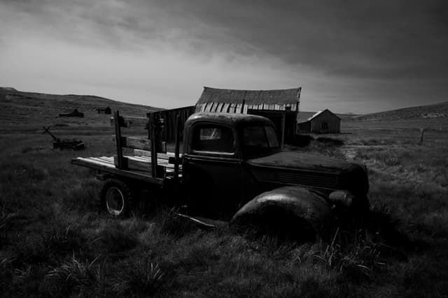 A black and white scene featuring an old, abandoned truck in a grassy field with a rustic barn and distant hills under a cloudy sky