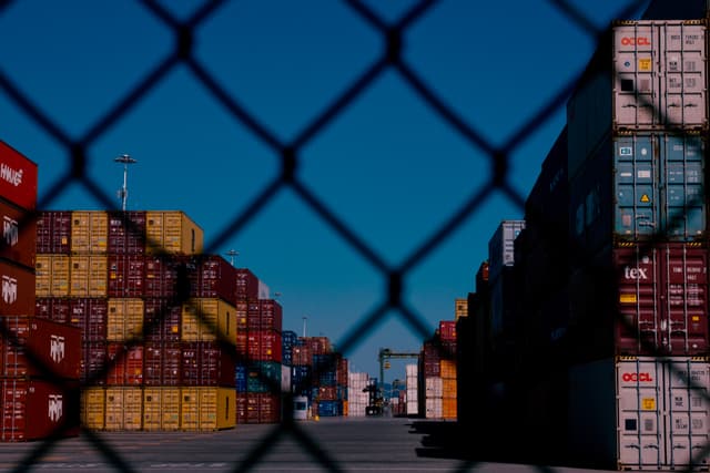 Stacks of colorful shipping containers are visible through a chain-link fence under a clear blue sky
