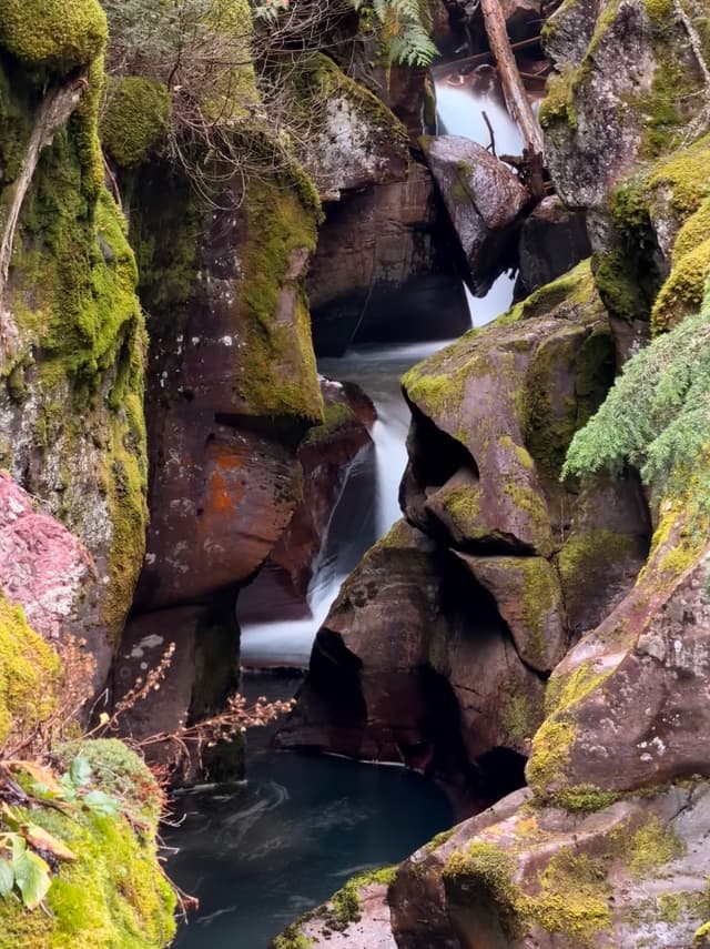 A small waterfall threads through a narrow, moss-covered rocky gorge, pooling in a blue basin below