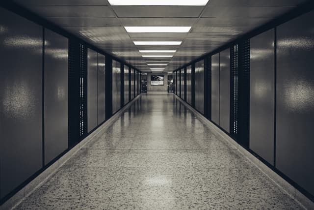 A long, empty corridor with a polished floor, flanked by dark walls and illuminated by overhead fluorescent lights