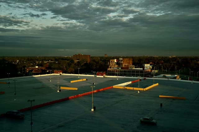 Rooftop parking deck at dusk, yellow barriers glowing in low sunlight, with a cityscape and dramatic clouds in the background