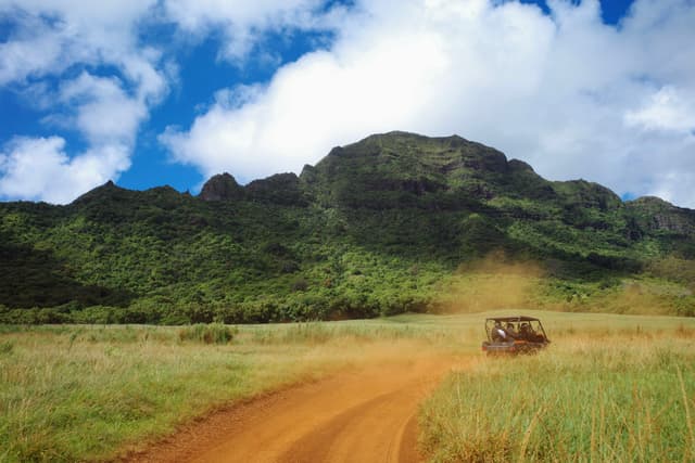 A rugged vehicle drives along a dirt road through a grassy landscape, with a lush green mountain and a partly cloudy sky in the background
