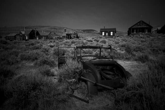 A black and white scene of an abandoned, overgrown ghost town with dilapidated buildings and a rusted, broken-down car in the foreground