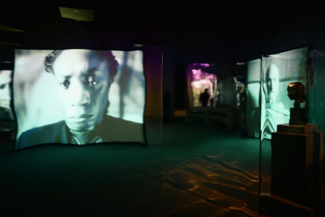 A dimly lit exhibition space with large screens displaying black and white portraits, creating an immersive atmosphere