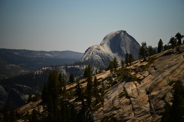 A mountainous landscape with a prominent granite dome in the background, surrounded by trees and rocky terrain under a clear sky