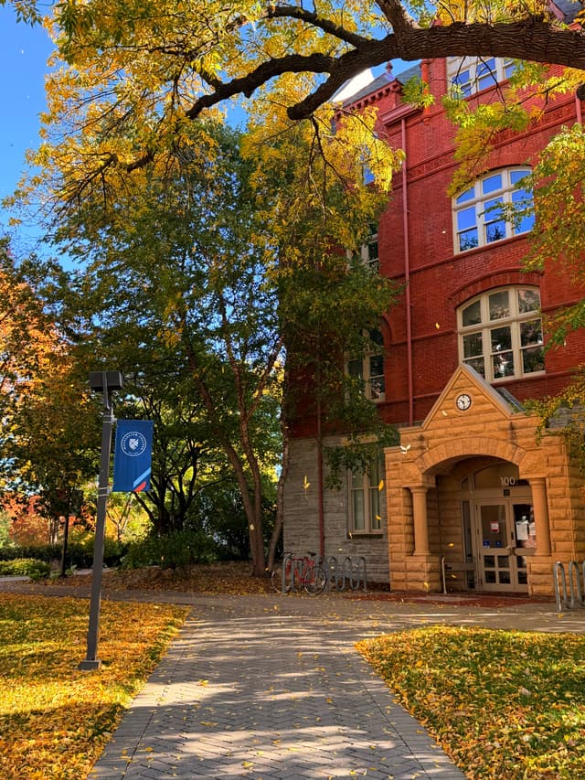 Red-brick building with an arched doorway, autumn foliage, and a paved walkway along a leaf-strewn path