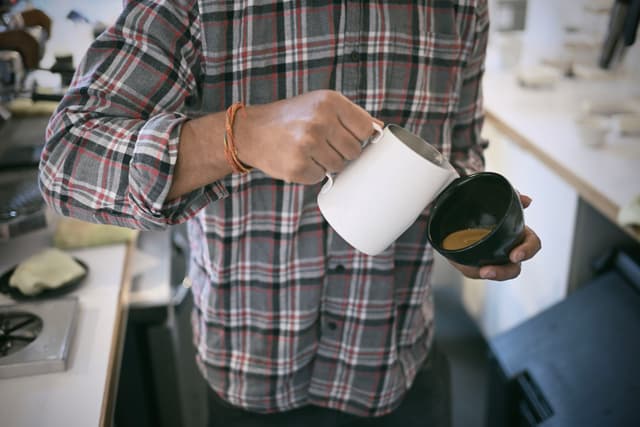 A person in a plaid shirt pouring milk from a white pitcher into a cup of coffee