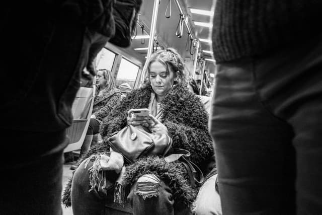 A person sitting on public transportation, focused on their phone, surrounded by other passengers. The scene is in black and white