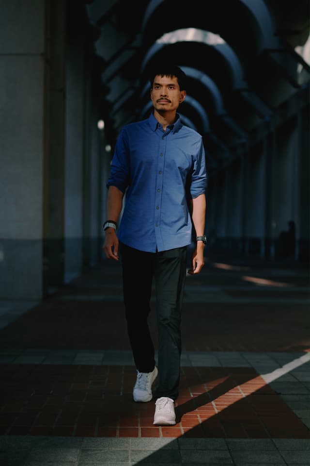 A man walks confidently through a shaded archway corridor, dressed in a blue shirt and dark pants, illuminated by natural light