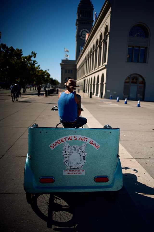 A person riding a pedicab along a street with a large building and clock tower in the background