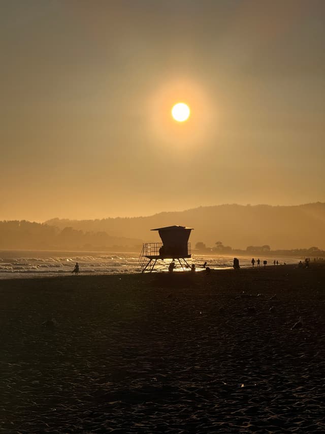 A lifeguard tower silhouetted against a sunset on a beach, with the sun low in the sky and hills in the background