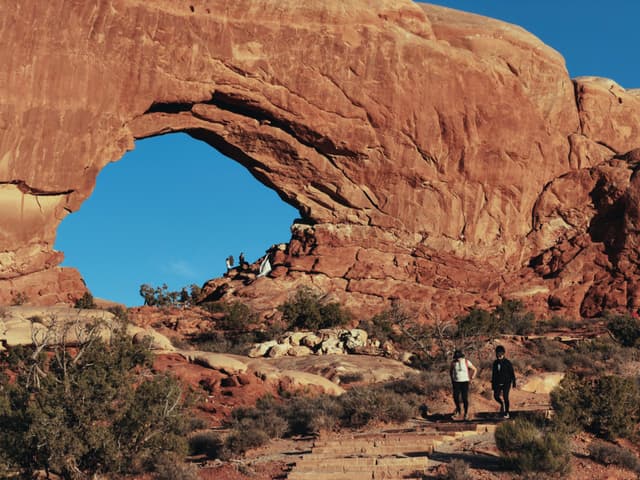 Hikers approach a large natural rock arch in a desert landscape under a clear blue sky