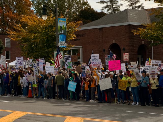 A large crowd lines a sidewalk in front of a brick building, holding protest signs and American flags on an autumn day