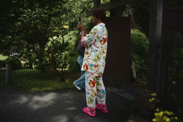 A person wearing colorful floral pajamas and pink shoes stands outdoors, holding a camera. The background features greenery and a wooden structure