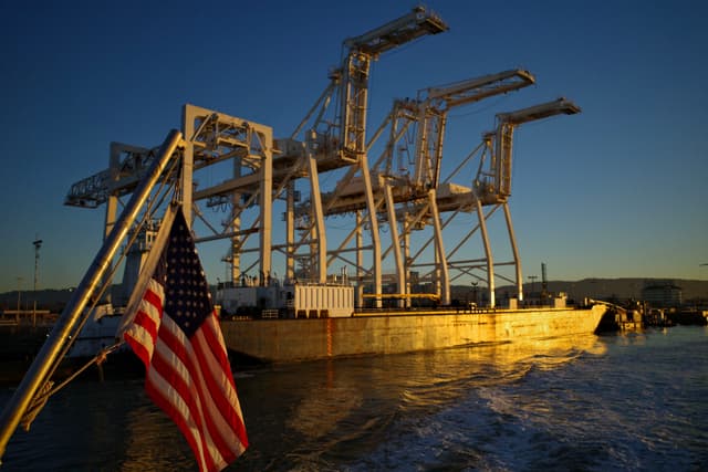 A group of large industrial cranes on a dock, with an American flag in the foreground and water reflecting the golden light of sunset