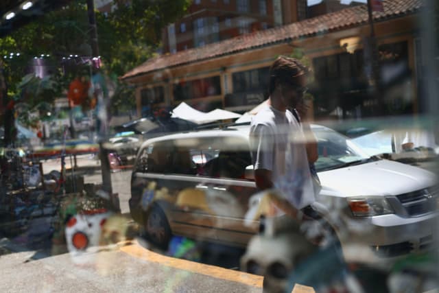 A person walking past a parked car on a sunny street, with reflections of various objects in a window