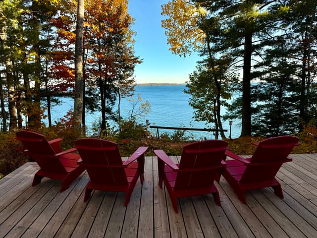 Four red Adirondack chairs on a wooden deck facing a calm lake, framed by tall trees with early autumn foliage