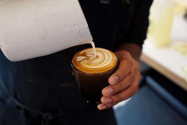A person pouring milk into a cup of coffee, creating latte art with a leaf design