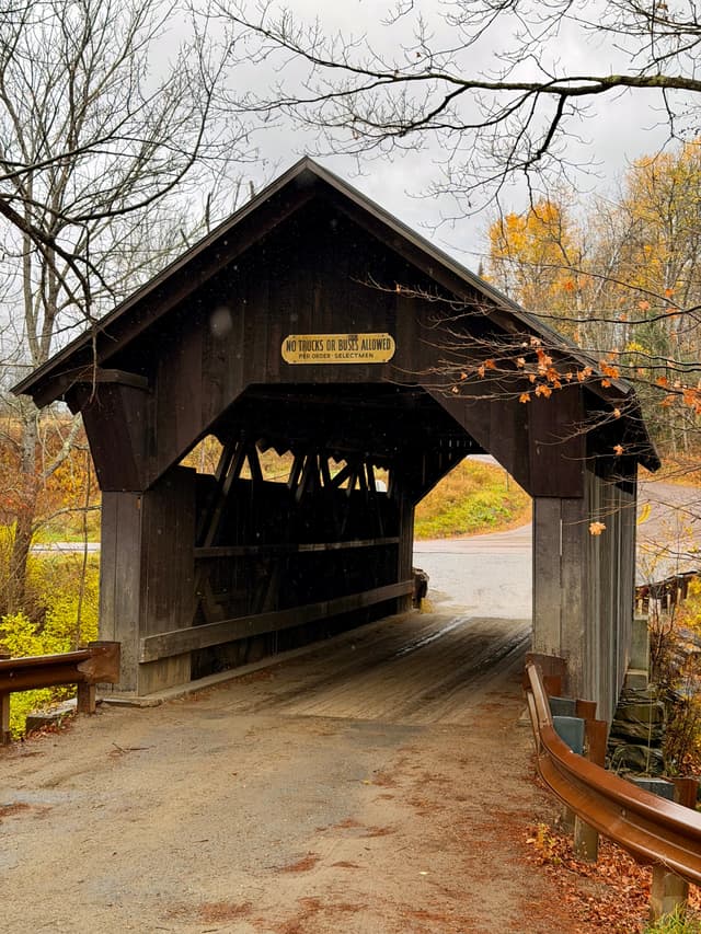 Rustic wooden covered bridge on a rainy autumn day, with wet road and colorful foliage in the background
