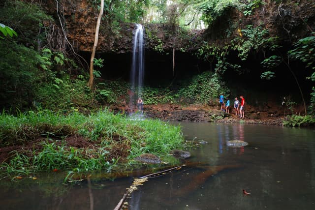A small waterfall cascades into a tranquil pool surrounded by lush greenery, with three people standing near the water's edge
