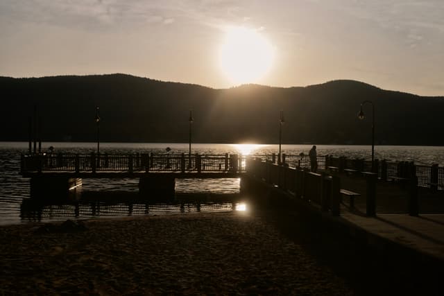 Sunset over a lakeside pier with silhouetted people and lampposts, mountains in the background, and sunlight reflecting on the water