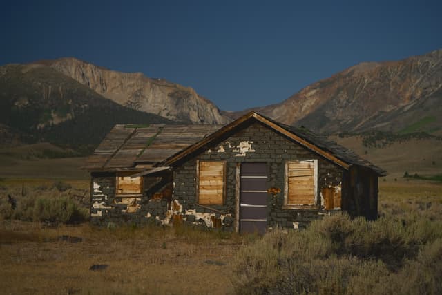 An abandoned, weathered cabin with boarded-up windows stands in a grassy field, surrounded by mountains under a clear blue sky